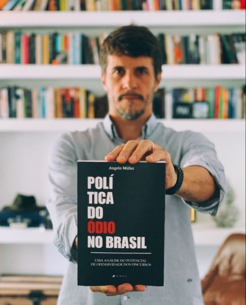 Angelo Muller, doutor em Comunicação Politica pela Famecos, exibindo seu livro Politica do Ódio no Brasil. Ele aparece atrás desfocado e seu livro aparece à frente, com capa negra, letras brancas (exceto por ódio, que está em vermelho)
